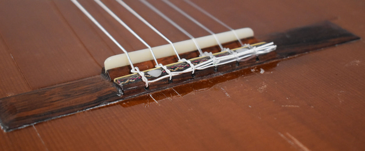 Close-up of the classical guitar's bridge and saddle, showing white nylon strings, a dark wood bridge, and wear on the natural finish.