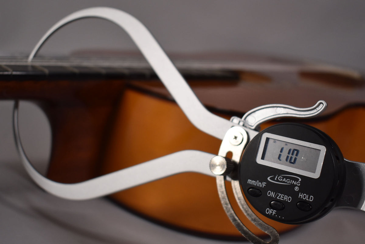 A digital caliper measures the string height of the natural finish classical guitar, showing "1.10" on its display.