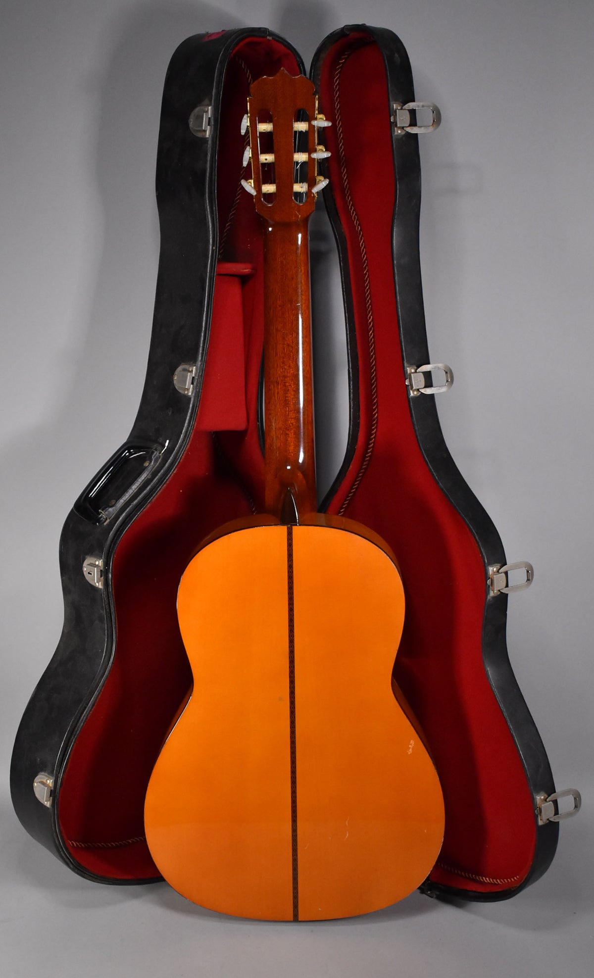 The classical guitar's glossy natural finish back and neck are visible, standing in a black hard case with red velvet lining.