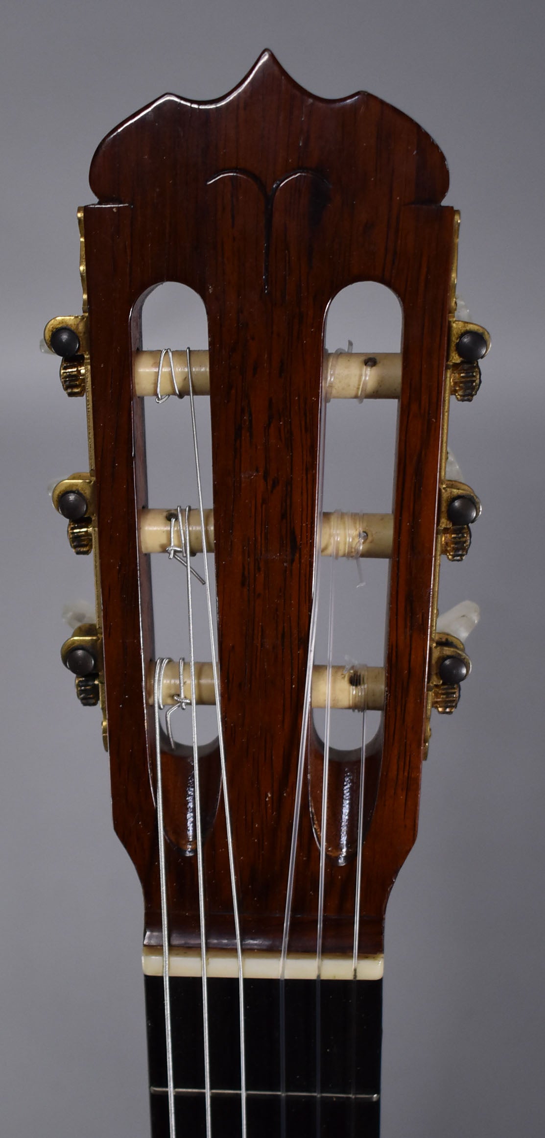 The dark brown headstock, with a decorative top edge, features gold-toned tuners with white buttons and nylon strings.