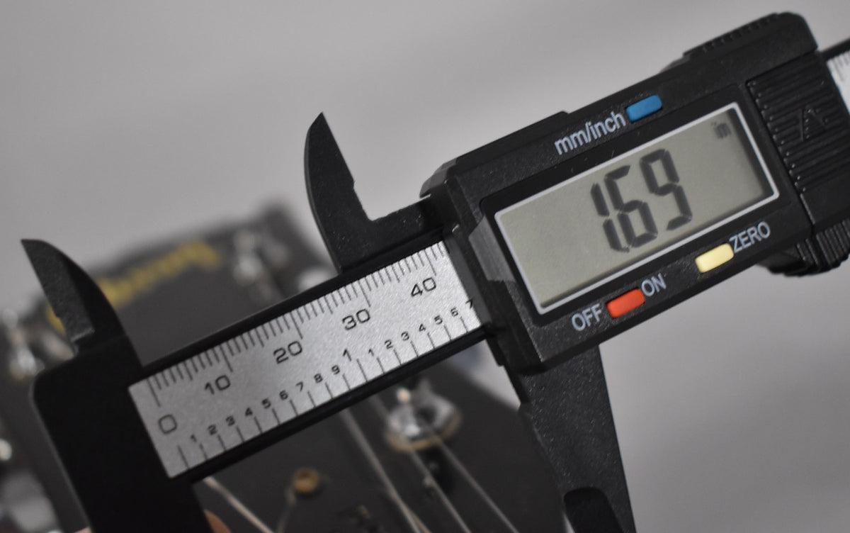 A digital caliper measures the guitar's nut width at 1.69 inches, with the dark headstock and tuning pegs blurred in the background.