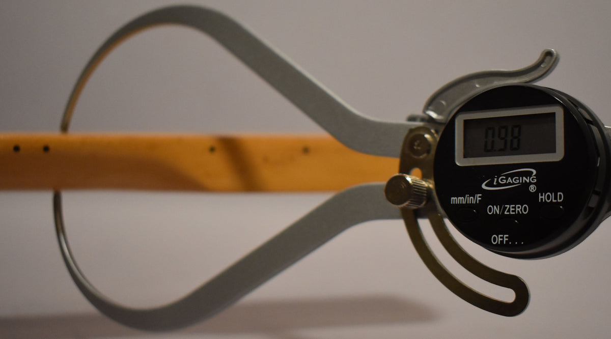 A digital caliper measures the maple neck's thickness at 0.98, showing the light-colored wood and side dot markers.