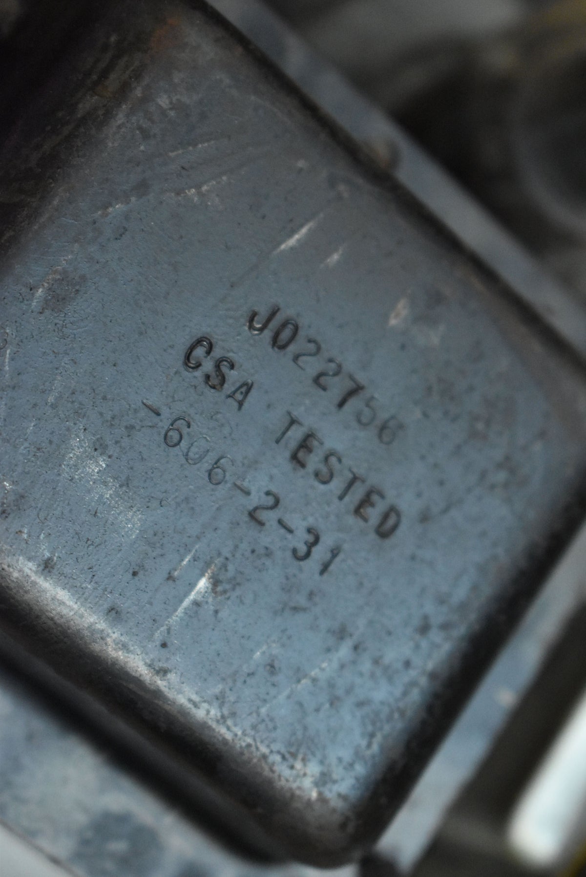 Close-up of a dark gray metal component with stamped text "J022756 CSA TESTED -606-2-31" and visible wear.