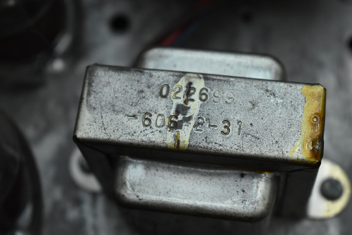 Close-up of a tarnished, rectangular silver metal component with stamped numbers and a worn yellow sticker, showing rust and grime.