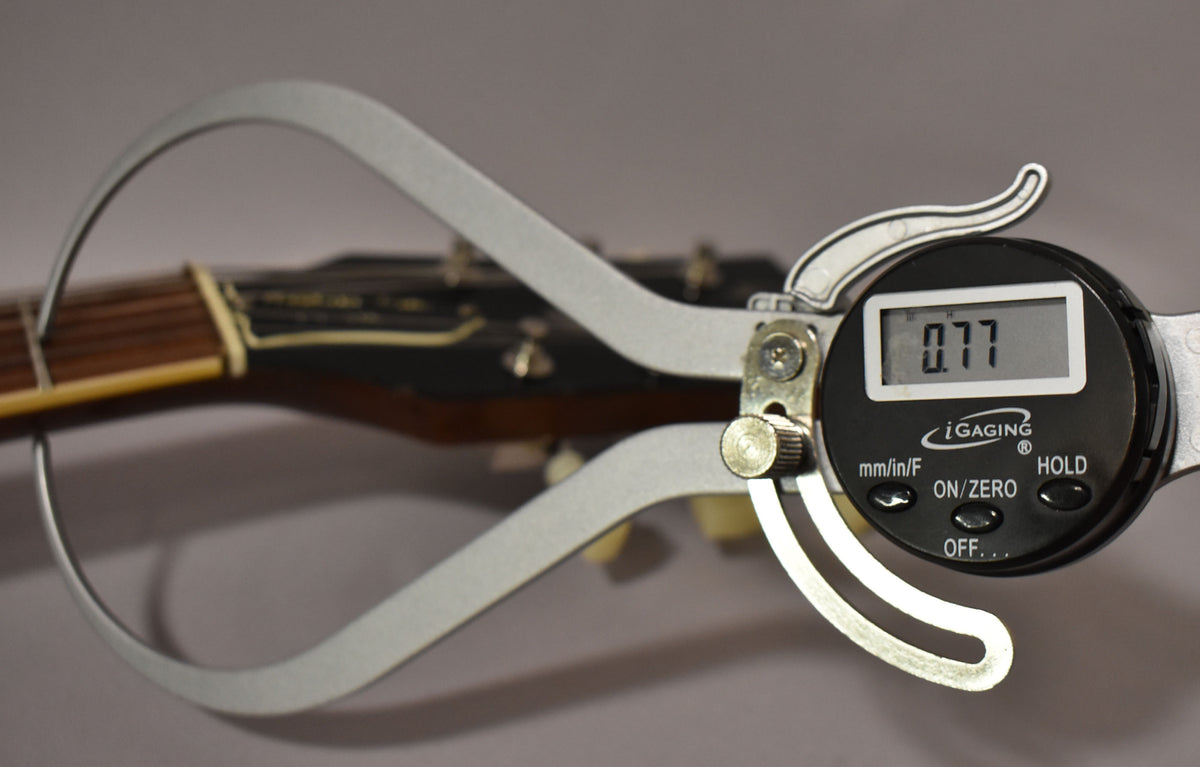 A digital caliper measures the guitar's nut width at 0.77 inches, with the dark headstock and fretboard blurred in the background.