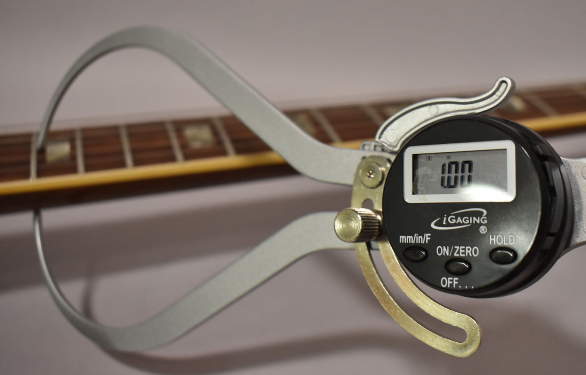 Close-up of a digital caliper measuring the guitar's fretboard, showing the dark wood, rectangular inlays, and binding.