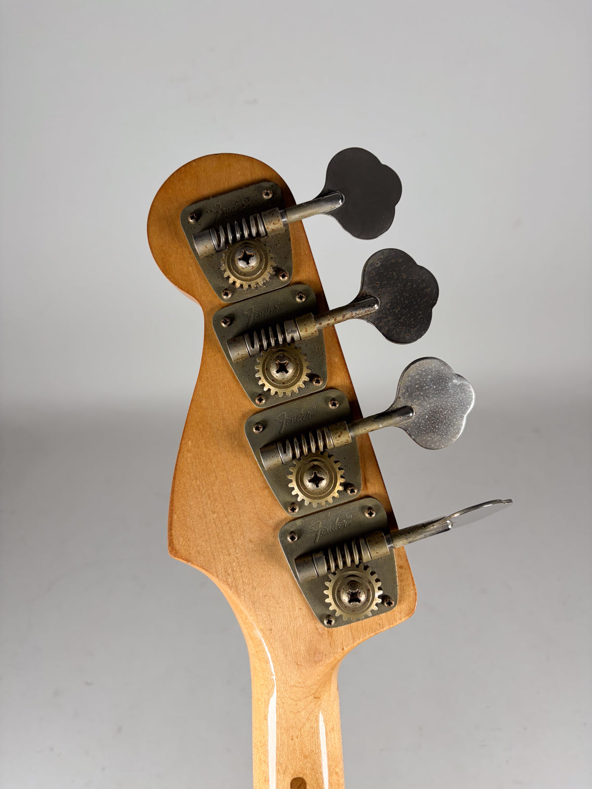 Close-up of the bass headstock from behind, showing four clover-leaf tuners with tarnished metal plates and gears on a light wood neck.