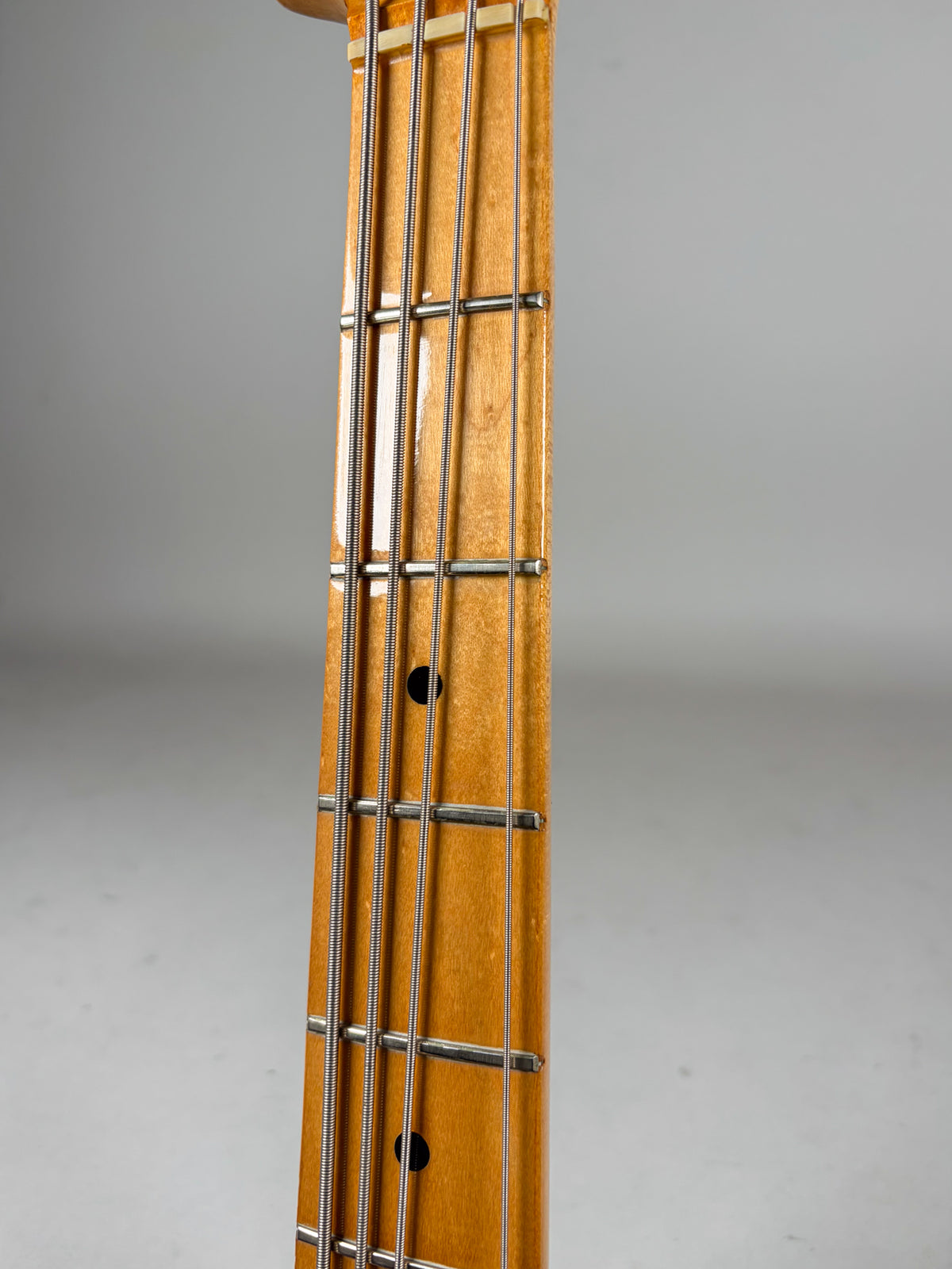 Close-up of the bass's maple fretboard, showing four strings, metal frets, and black dot inlays on a glossy, ambered finish.