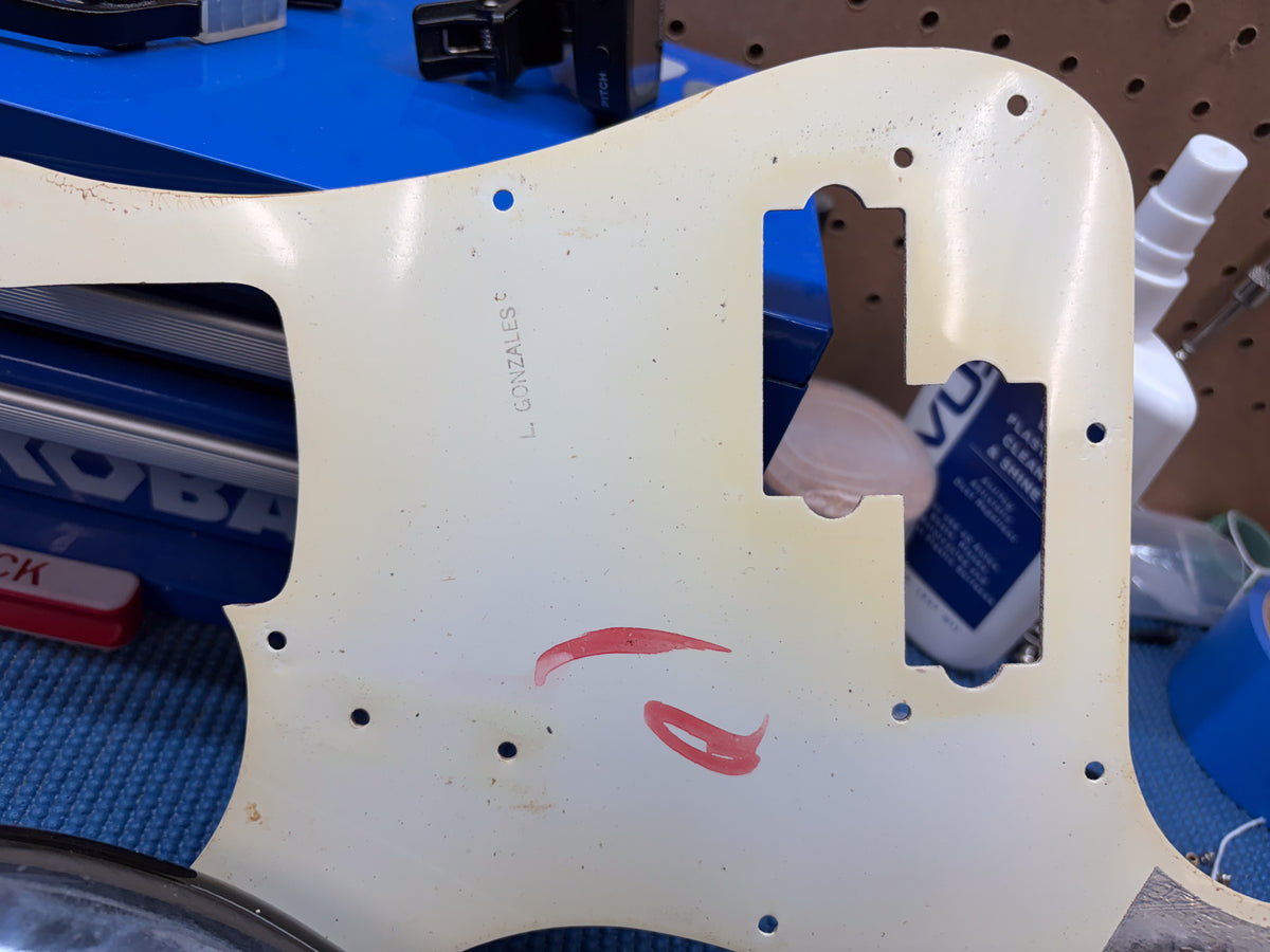 Close-up of the bass's aged cream pickguard, showing wear, some red marks, and "L. GONZALES" written on the back.