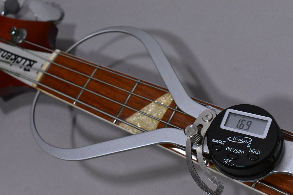 A digital caliper measures the bass's fretboard width at the nut, showing 1.69 inches, with pearloid triangle inlays visible.