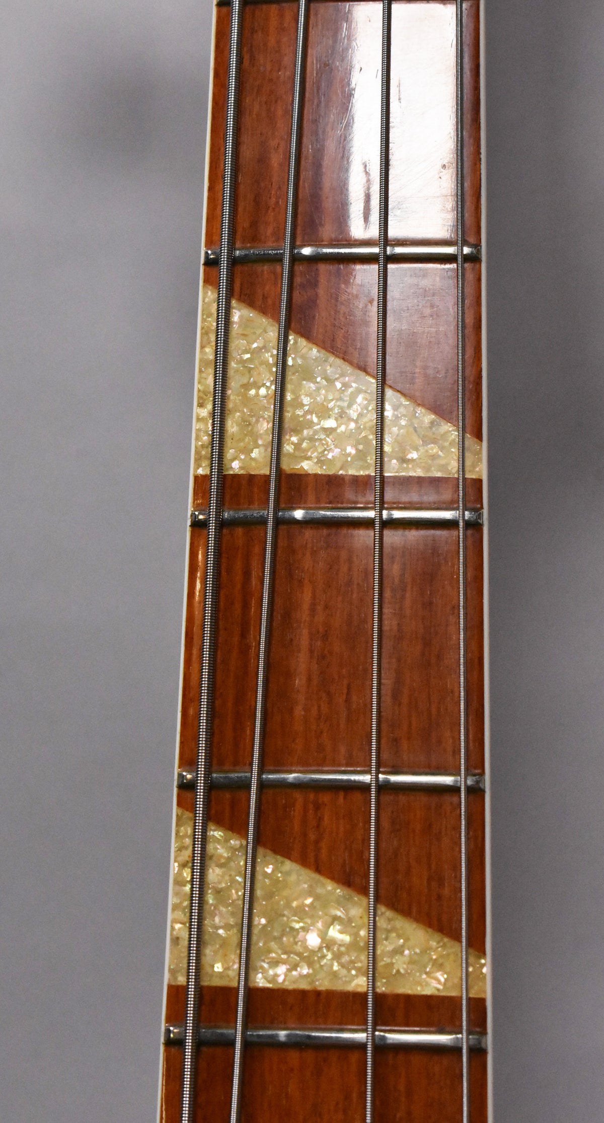 Close-up of the bass's dark brown fretboard with four strings, showing trapezoidal mother-of-pearl inlays and some wear on the frets.