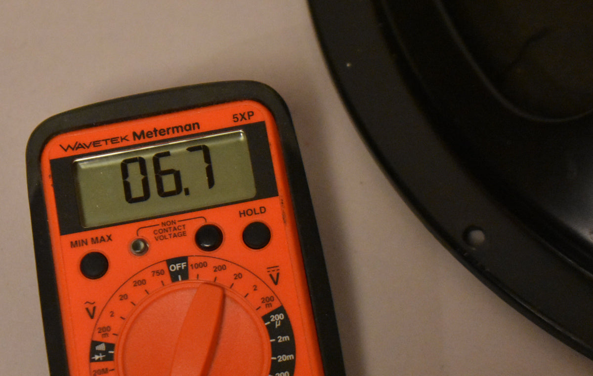 An orange multimeter displays "06.7" next to the edge of a black speaker frame with a mounting hole.
