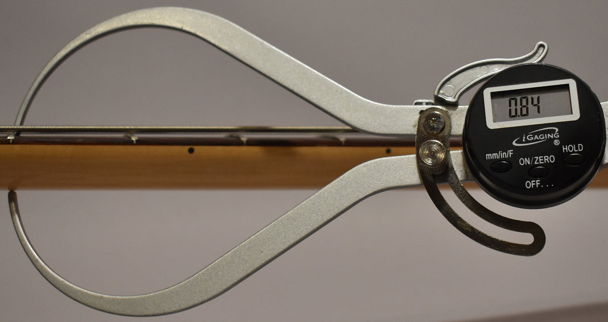 Digital calipers measure the bass's neck relief at 0.84, with the light maple neck and fretboard visible.