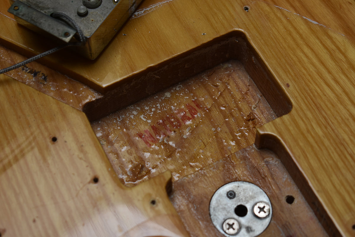 Close-up of a guitar body's neck pocket with a faint red stamp, a metal plate with three screws, and a pickup in the upper left.