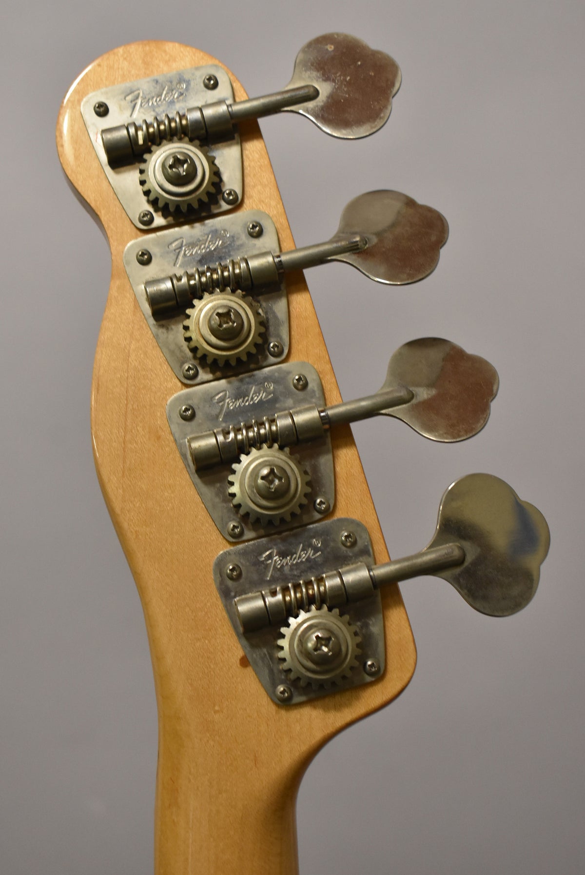 Close-up of the bass headstock from behind, showing four tarnished open-gear tuners on a light natural wood finish.