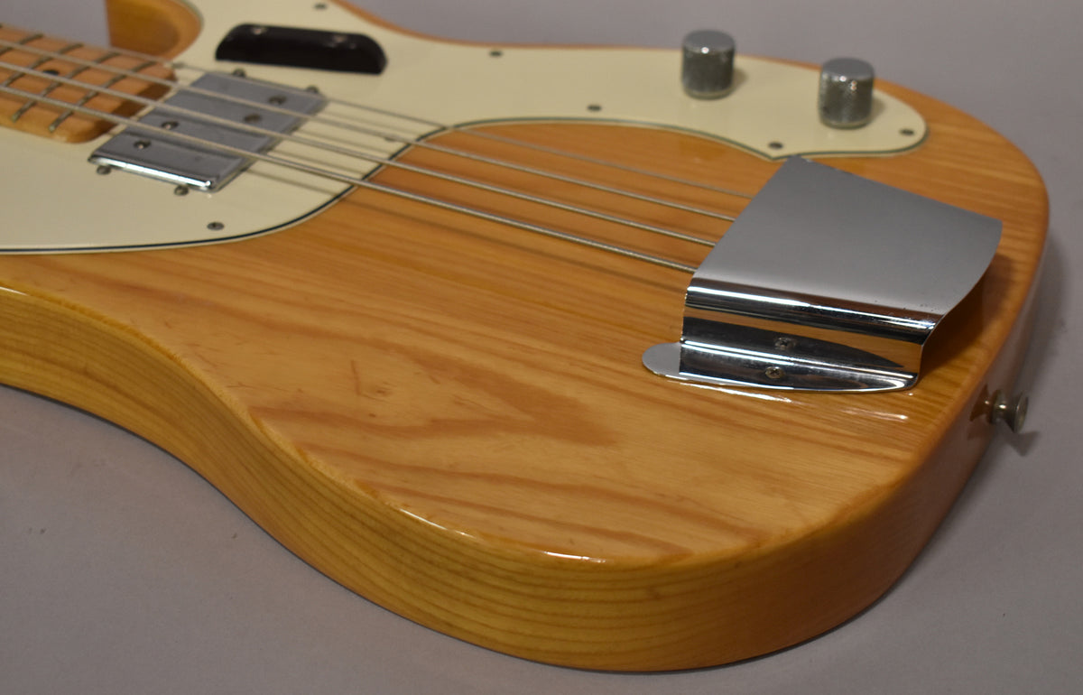 Close-up of the bass body, showing the natural ash grain, cream pickguard, chrome bridge cover, and two control knobs.