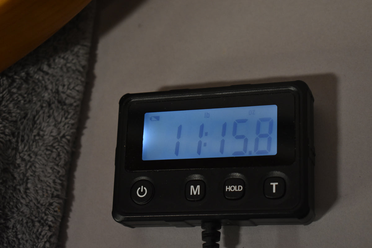 Close-up of a digital scale displaying "11:15.8" in blue, with "lb" and "oz" indicators, and power, M, HOLD, and T buttons.
