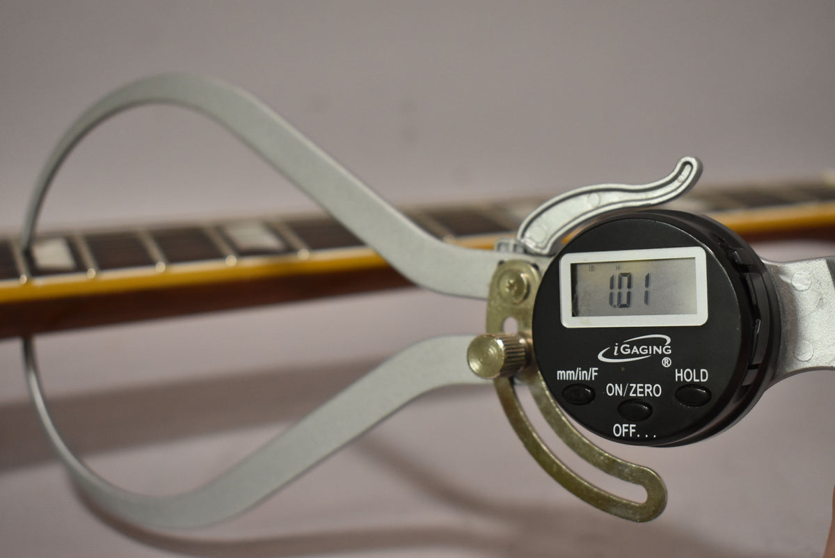 A digital caliper measures the guitar's fretboard, showing "1.01" on its display, with block inlays visible.
