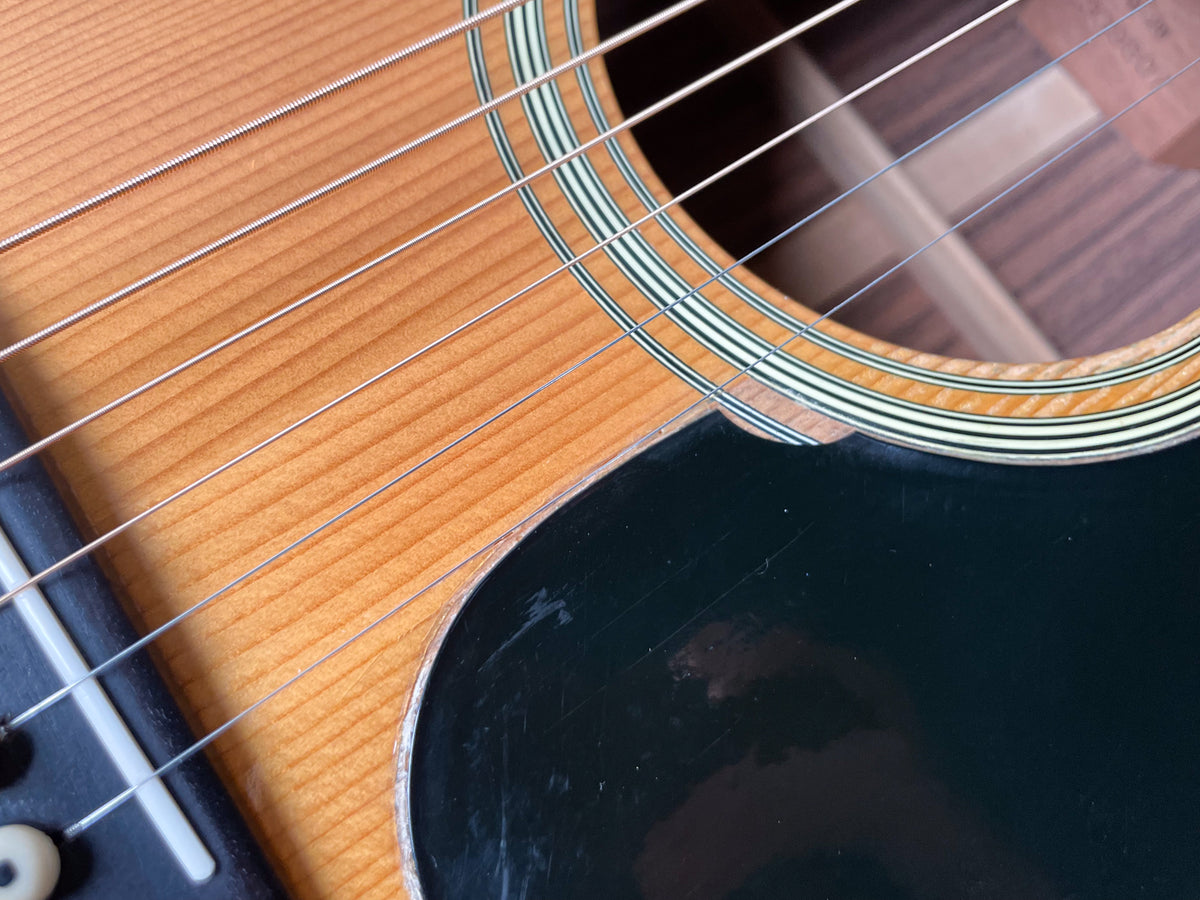 Close-up of the guitar's body, showing the light wood top, soundhole rosette, black pickguard with scratches, and six strings.