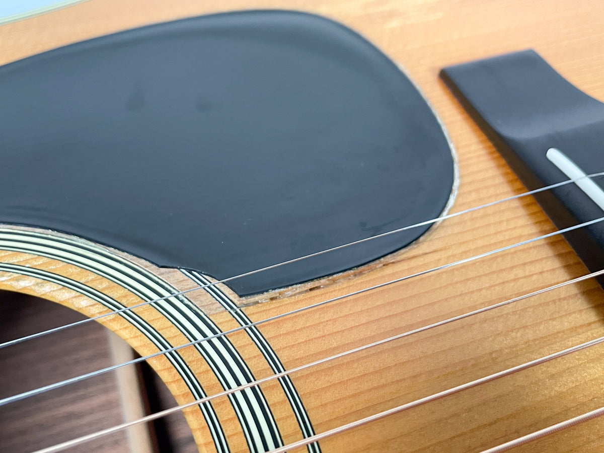 Close-up of the guitar's natural wood top, showing the black pickguard, soundhole rosette, bridge, and six strings.