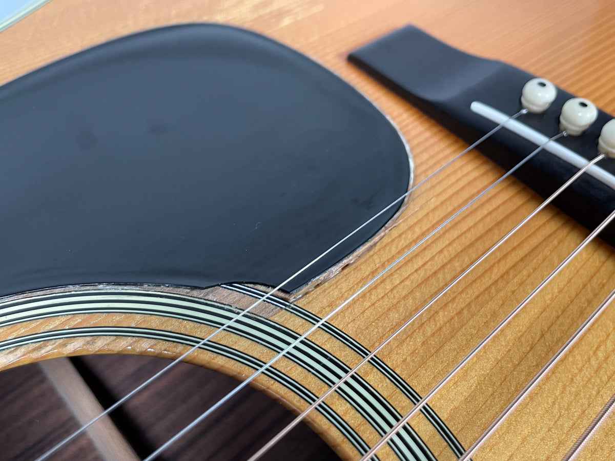 Close-up of the guitar's natural finish soundboard, showing the black pickguard lifting near the soundhole and bridge.