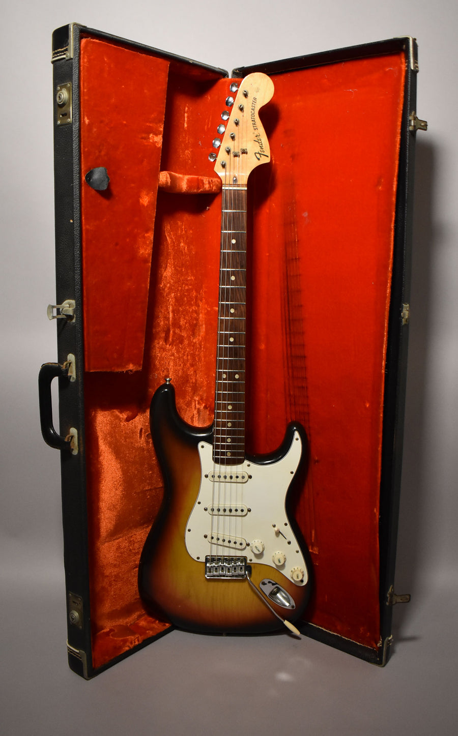Full-length sunburst electric guitar with white pickguard and three single-coil pickups, resting in an open black hard case with red plush lining.