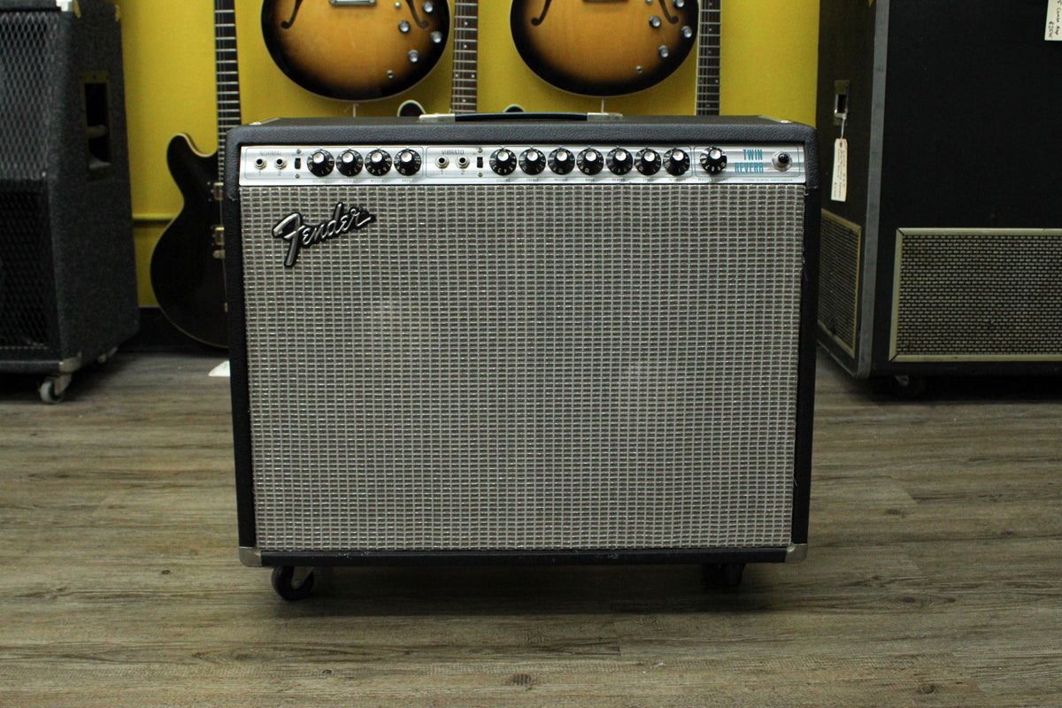 Front view of a black amp with a silver grille, black control panel with 12 knobs, and four casters.