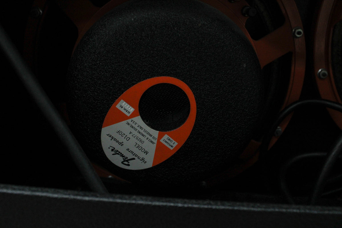 Close-up of a black speaker cone with an orange and white oval label, revealing part of a second speaker and orange speaker frames.
