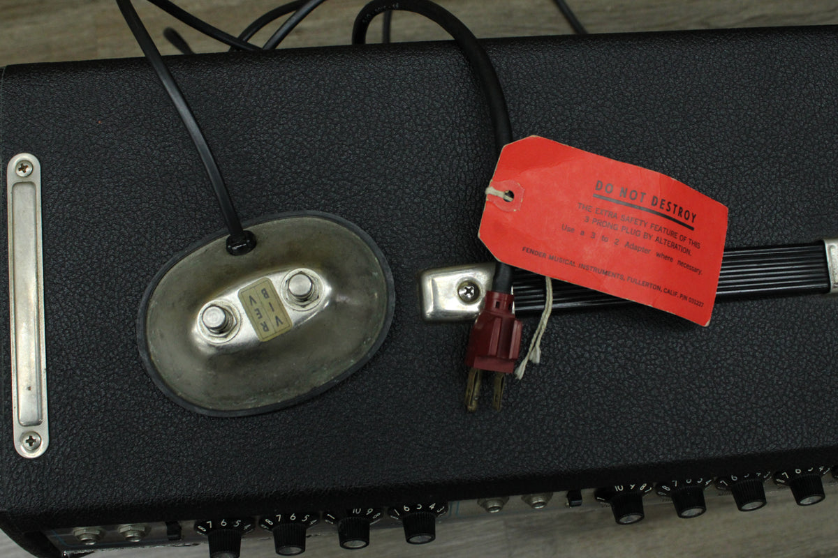 Top-down view of the black amp, showing a silver footswitch, a red warning tag, and several black control knobs.