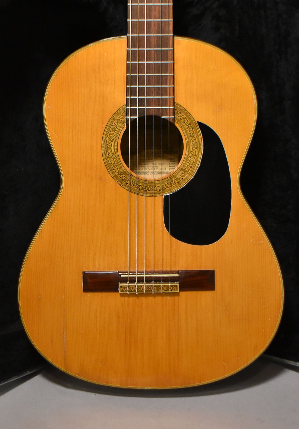 Full front view of a light-wood classical guitar with a dark pickguard, ornate rosette, and some finish wear on the body.