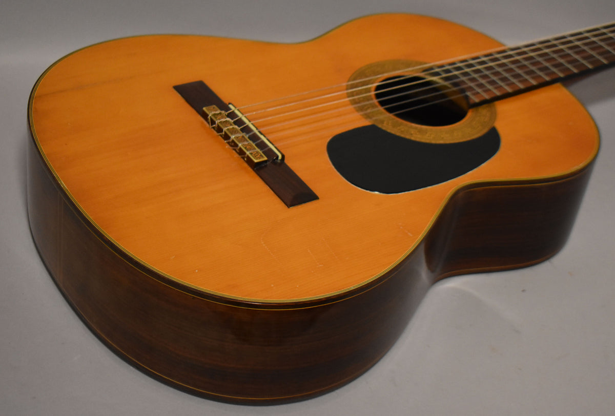 The classical guitar's light-colored top with a black pickguard and dark sides is angled from the lower bout, showing light wear.