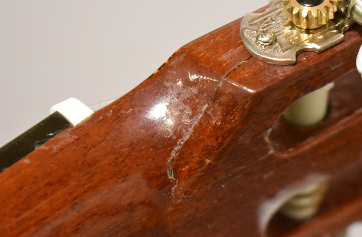 Close-up of the classical guitar's headstock, showing finish wear and checking on the dark brown wood, with a gold and silver tuner.