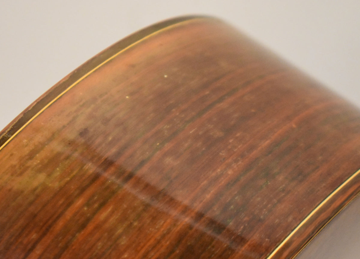 Close-up of the guitar's dark reddish-brown wood side with a glossy finish, showing wood grain, light wear, and a thin gold binding.