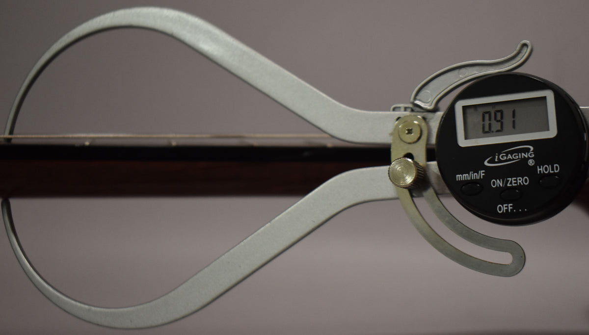 A digital caliper measures the guitar's dark brown fretboard, showing a reading of 0.91 on its black display.