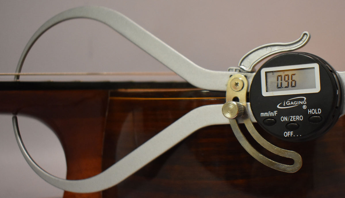 A digital caliper measures the string height at the 12th fret of the dark brown classical guitar, displaying "0.96".