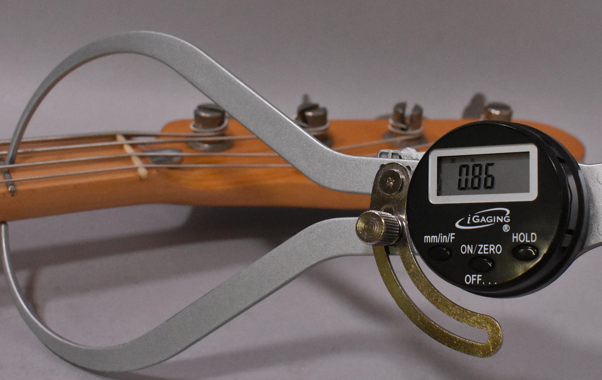 A digital caliper measures the string height on the bass's light-colored maple neck, showing "0.86" on its display.