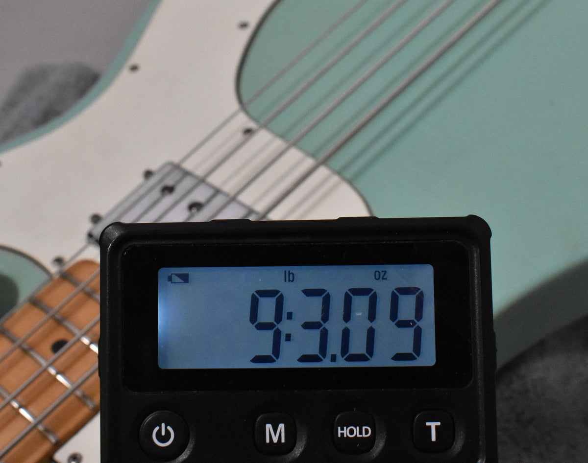 A digital scale displays "9:3.09 lb oz" in the foreground, with a seafoam green bass body and maple neck visible behind it.