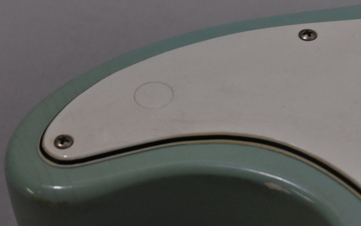 Close-up of the bass's light green body and white pickguard, showing finish checking, scratches, and a small circular mark.
