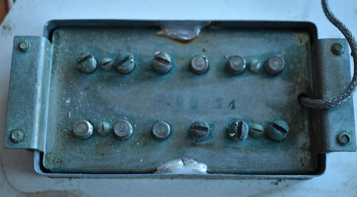Close-up of a tarnished metal humbucker pickup with two rows of six pole pieces, some slotted, some round, and a braided wire.