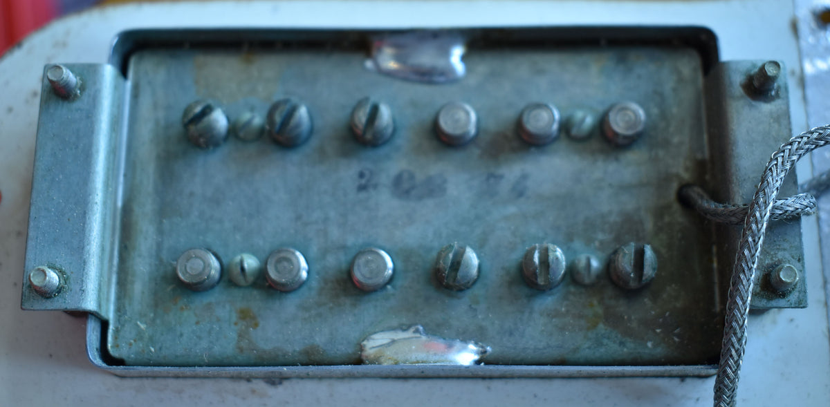 Close-up of a tarnished metal humbucker pickup with exposed pole pieces, a braided wire, and "2 08 74" stamped on it.