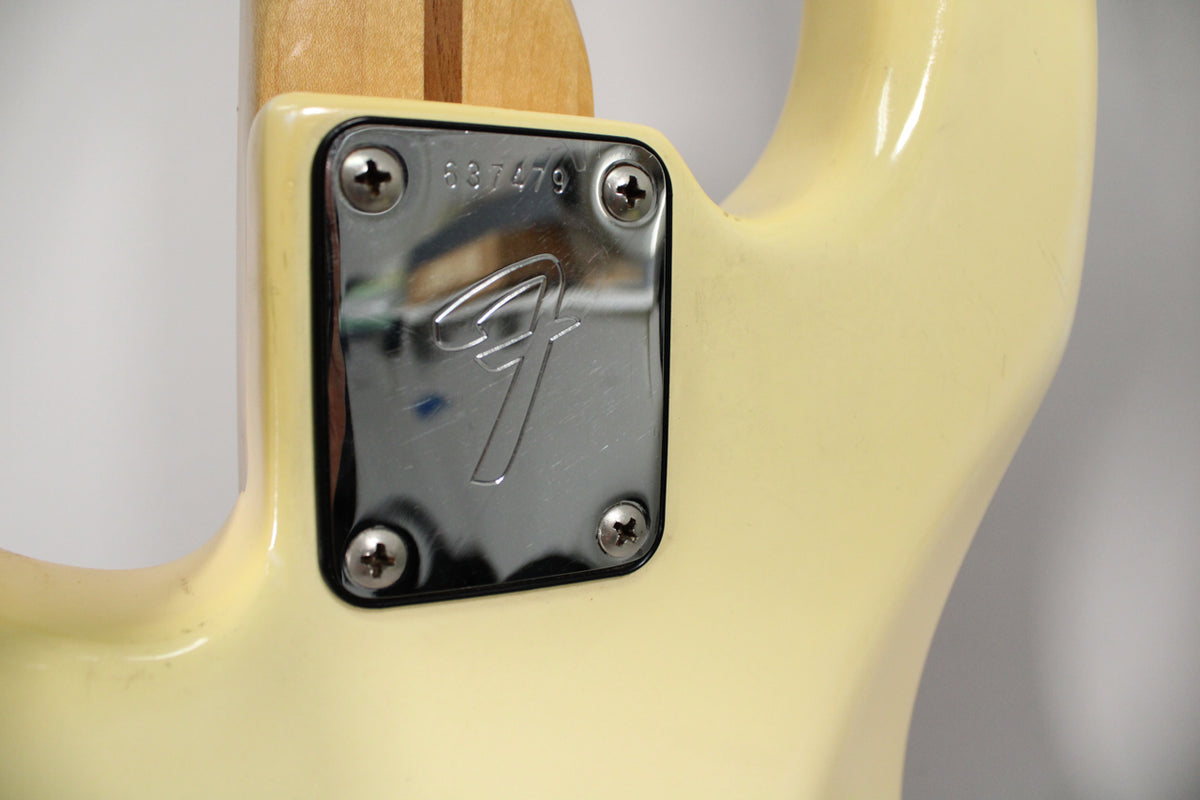 Close-up of the bass's neck plate, showing a chrome finish with a serial number and light scratches, bolted to the aged Olympic White body.