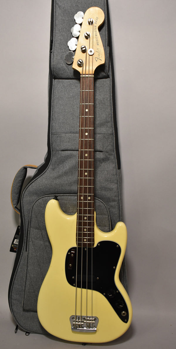 Full-length view of a creamy white bass with a black pickguard, rosewood fretboard, and a grey gig bag behind it.