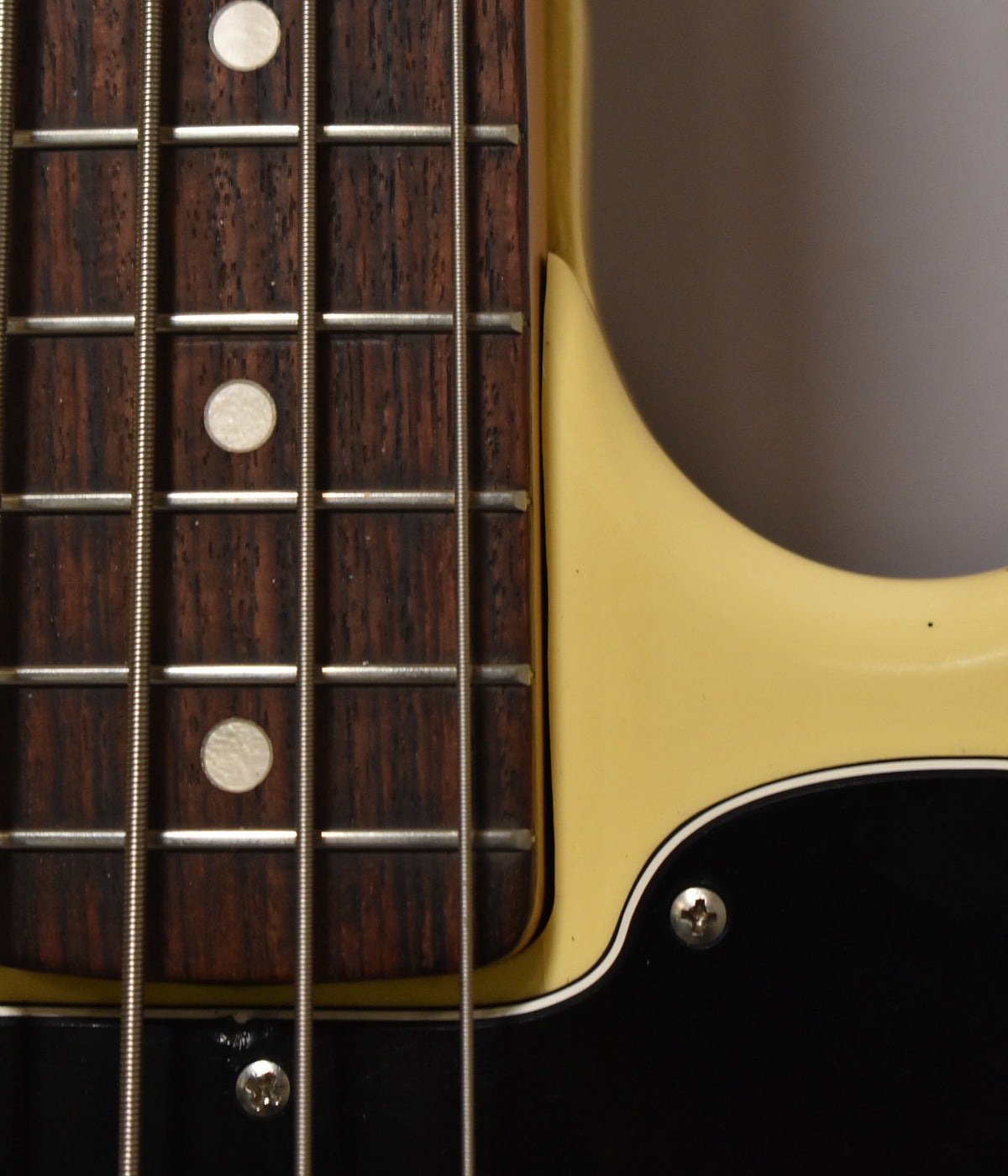 Close-up of the bass's dark fretboard with white dot inlays, showing light wear, next to the aged Olympic White body and black pickguard.