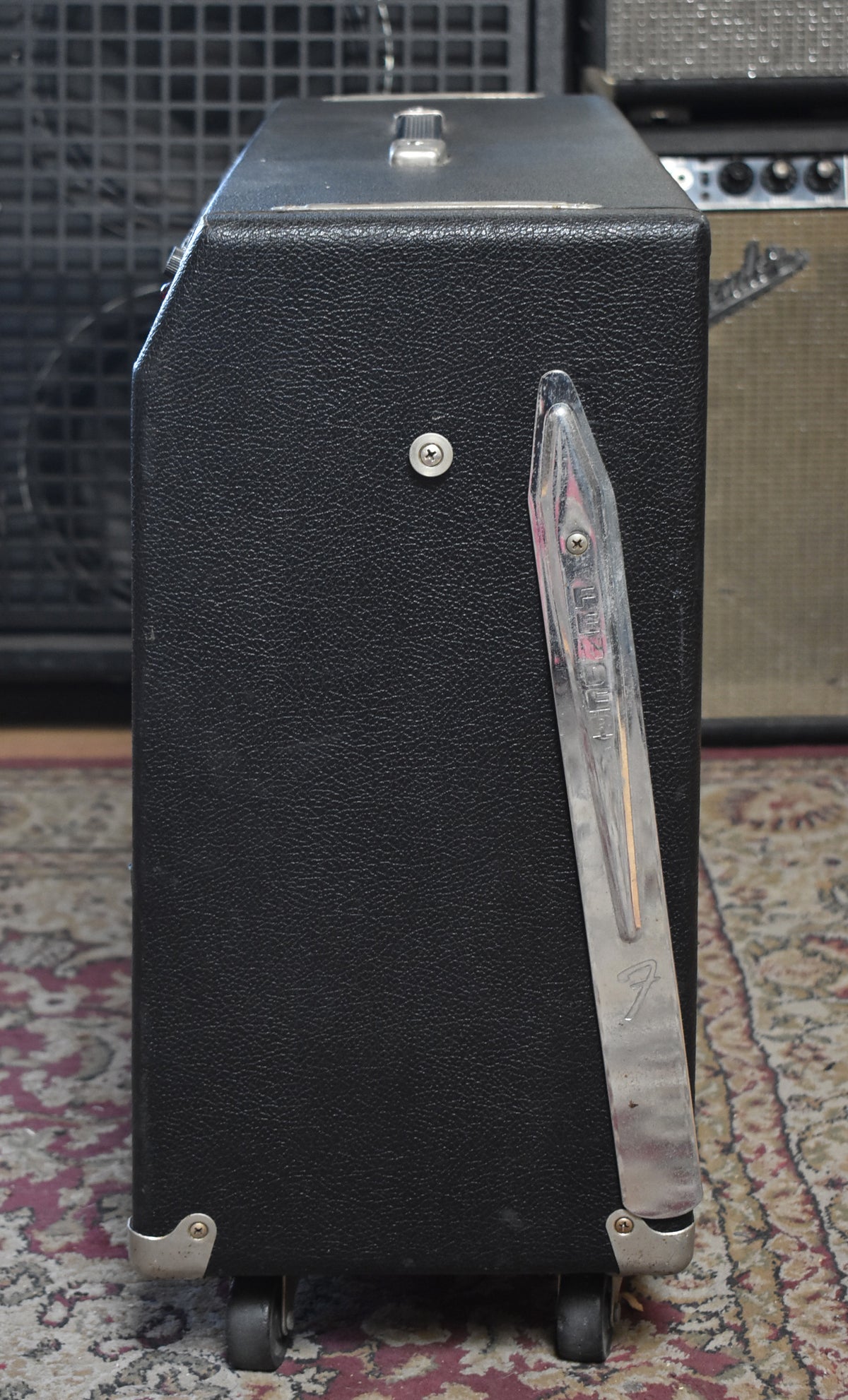 Right side view of the black tolex amp, showing a chrome tilt-back leg with a worn Fender logo, metal corner protectors, and casters.