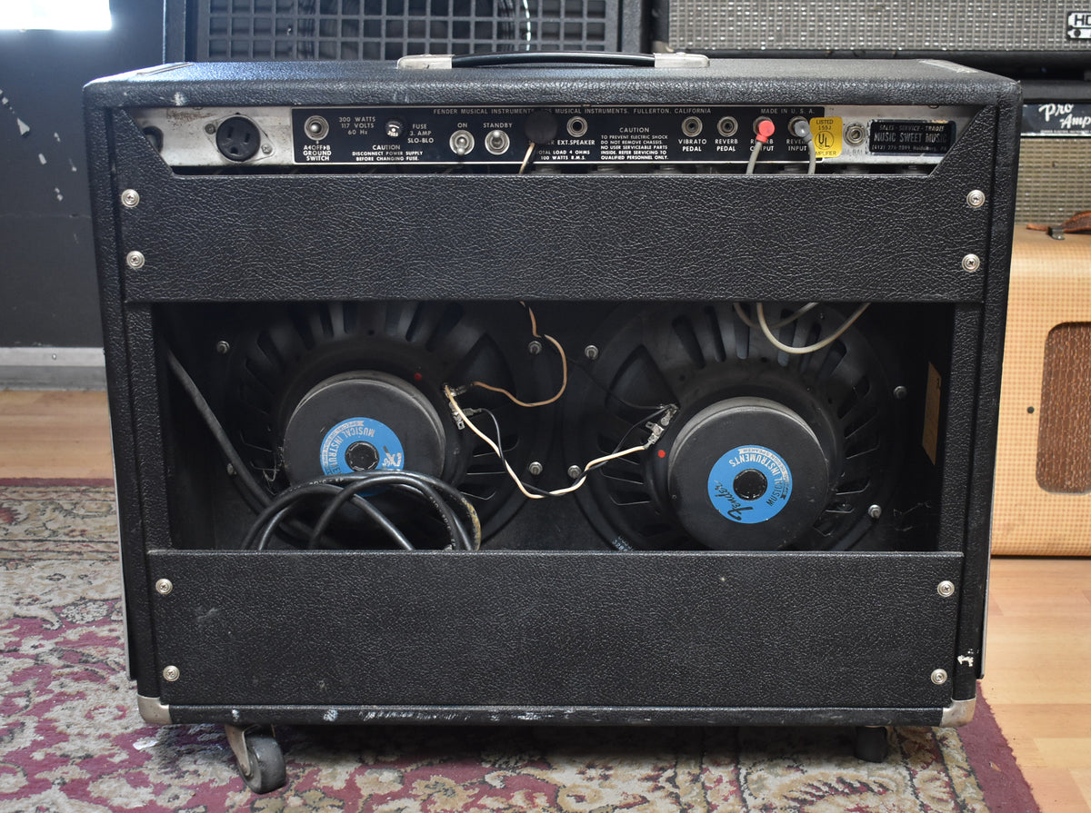 Rear view of the black textured amplifier, revealing two large speakers with blue labels, exposed wiring, and a metal control panel.