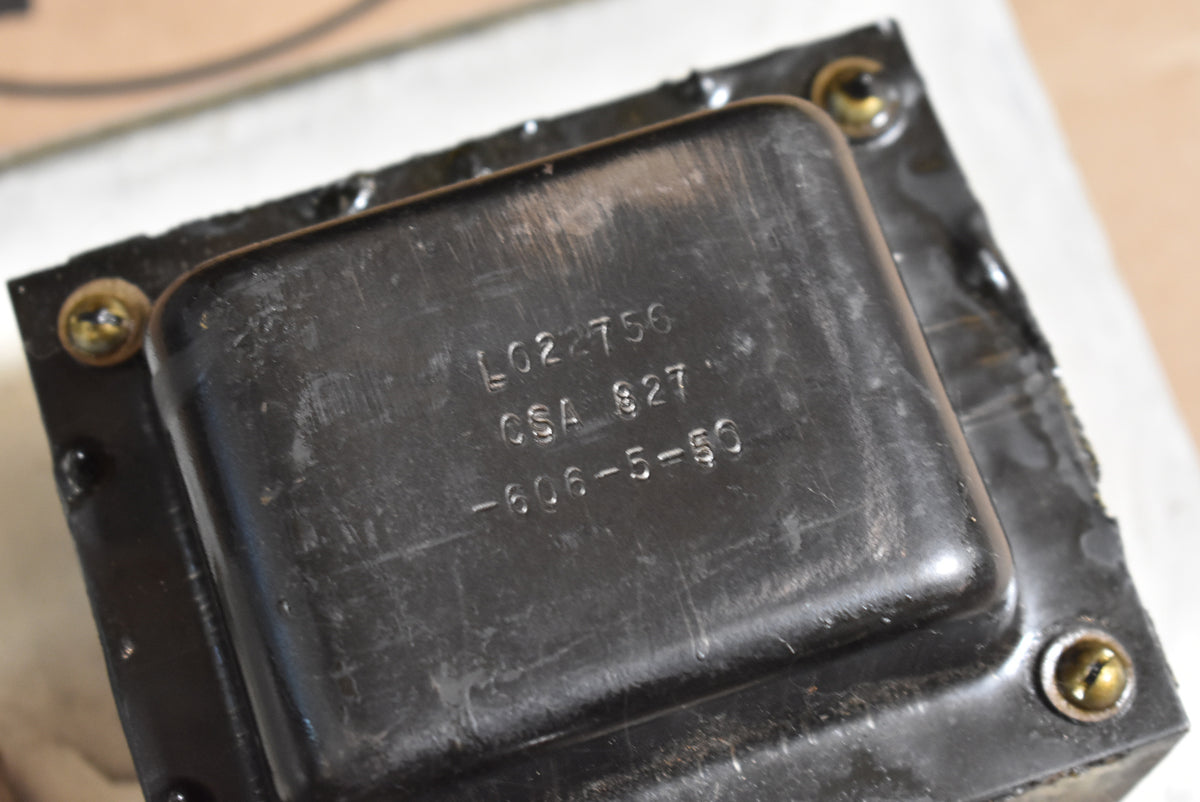 Close-up of a black metal transformer with visible wear, mounted with four brass screws, showing stamped numbers on top.
