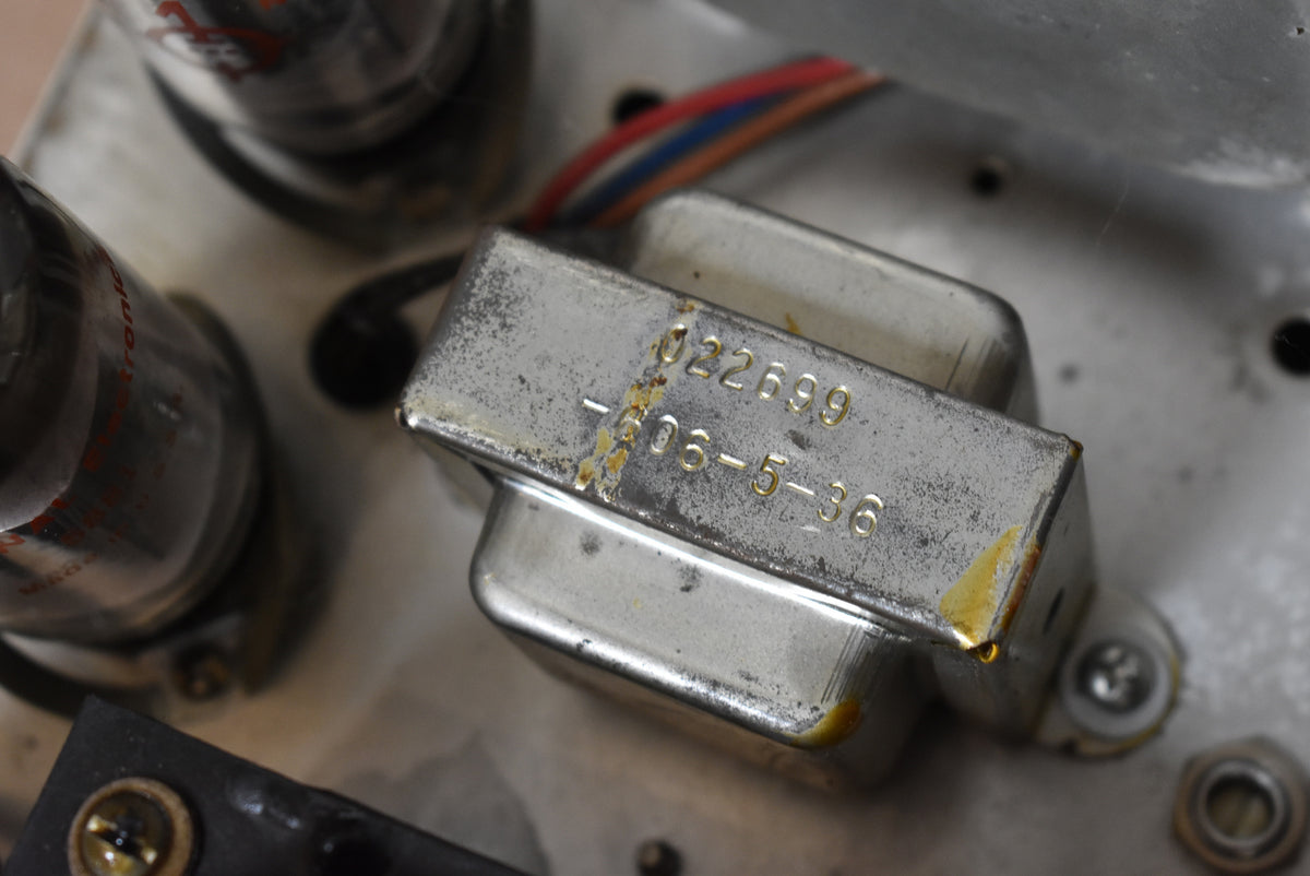 Close-up of the amp's internal components, showing a tarnished silver transformer with stamped numbers and visible rust spots, next to vacuum tubes.