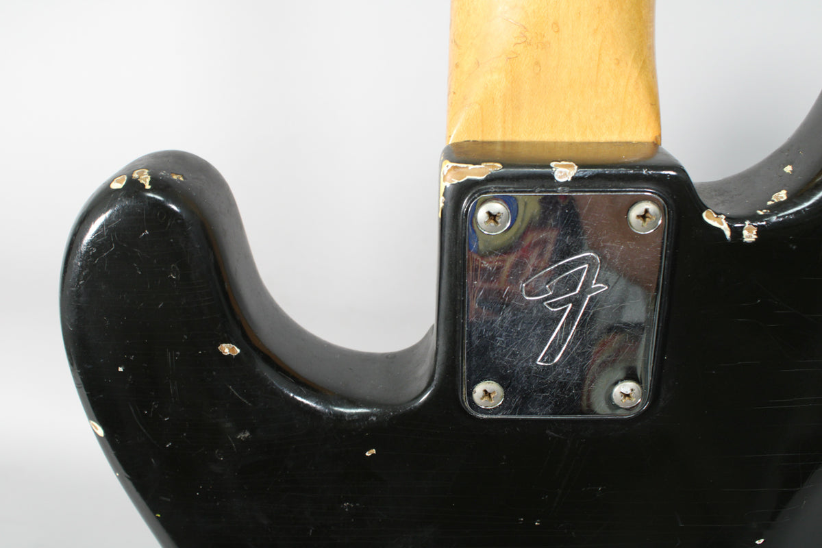 Close-up of the black bass's neck joint, showing a chrome neck plate with a script logo and significant paint chipping on the body.
