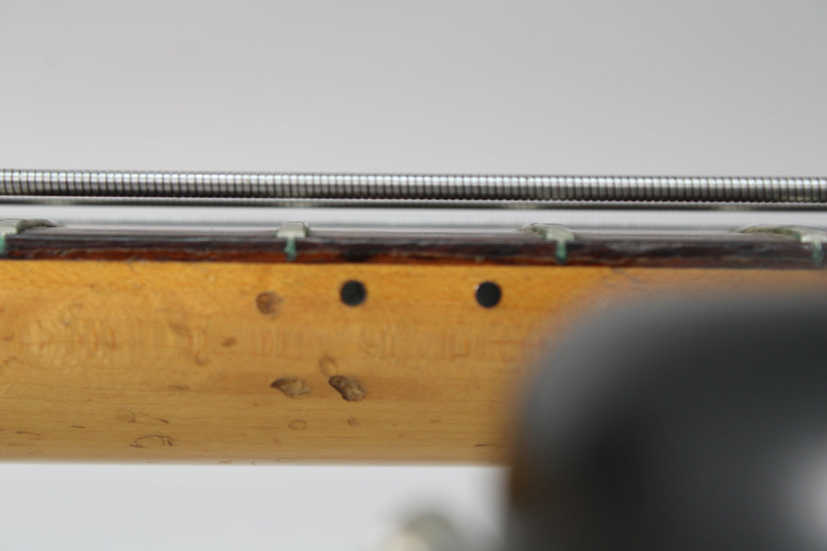 Close-up of the bass neck's side, showing the maple finish with dark circular fret markers and several dings and wear marks.