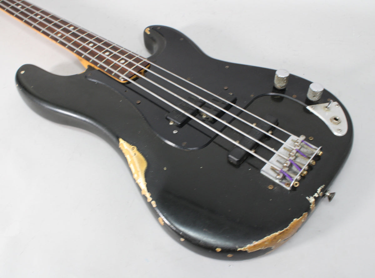 Black bass guitar body, heavily worn with paint chips revealing wood, angled from neck to bridge, showing P/J pickups and chrome controls.